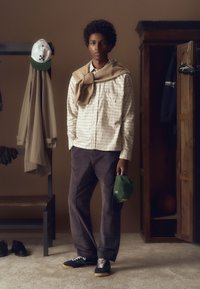 Person, der ein helles karriertes Hemd, einen braunen Pullover über den Schultern, dunkle Hosen und eine grüne Baseballkappe trägt. Im Hintergrund ist ein Garderobenständer zu sehen.