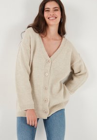 Cardigan lavorato a maglia beige con scollo a V profondo, sei bottoni sul davanti, polsini a coste e una vestibilità ampia. Indossato sopra jeans in denim.