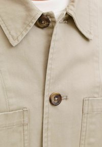 Χακί μπουφάν με μυτερό γιακά, δύο τσέπες στο στήθος και μεγάλο καφέ κουμπί. Το ύφασμα έχει απαλή υφή με ορατές λεπτομέρειες ραφής.