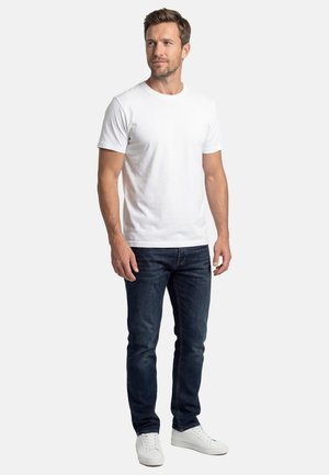 Uomo in piedi che indossa una maglietta bianca a maniche corte, jeans blu scuro e sneakers bianche, guarda leggermente di lato.