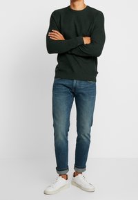 Dunkelgrüner Strickpullover mit Rundhalsausschnitt, kombiniert mit hellblauen Jeans und weißen Sneakers mit braunen Akzenten.