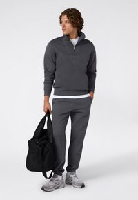 Uomo in felpa grigia scura con zip e pantaloni jogger abbinati, tiene una borsa da palestra nera, indossando scarpe da ginnastica grigie e bianche, guarda verso sinistra.