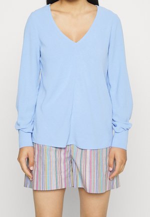 Persoon die een lichtblauw langermig V-hals shirt draagt en meerkleurige verticale gestreepte korte broek, staand met ontspannen handen langs het lichaam.