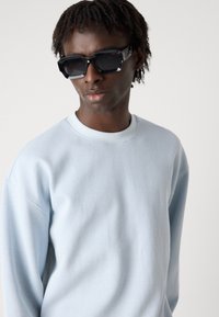Hanorac ribbed de culoare albastră deschisă, cu guler rotund. Ochelari de soare negri, cu un cadru unghiular și brațe laterale cu un model gri. Fără logo-uri vizibile.