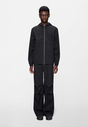 Νεαρός άνδρας που στέκεται ακίνητος, φοράει μαύρο φούτερ με κουκούλα και φερμουάρ, φαρδιά μαύρη παντελόνια και μαύρα παπούτσια, μπροστά σε απλό ανοιχτό φόντο.