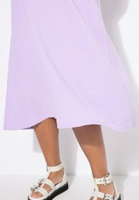 Lavendelfarbenes Textilkleid mit einem fließenden Saum, kombiniert mit weißen Sandalen mit Riemen, die Schnallenakzente und eine dicke schwarze Sohle haben.