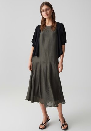 Frau trägt ein ärmelloses olivgrünes Midikleid mit einem schwarzen, kurzärmligen offenen Cardigan und schwarzen Riemchenabsätzen, steht vor einem einfarbigen Hintergrund.
