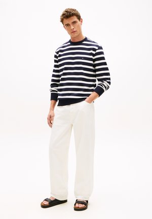 Młody mężczyzna ubrany w granatowo-białą koszulę w paski z długim rękawem, białe spodnie o szerokich nogawkach oraz czarne sandały, stoi z jedną ręką w kieszeni.