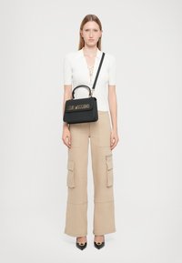 Black handbag with a top handle and shoulder strap, featuring gold "LOVE MOSCHINO" lettering. Model wears a fitted white top and beige cargo pants.