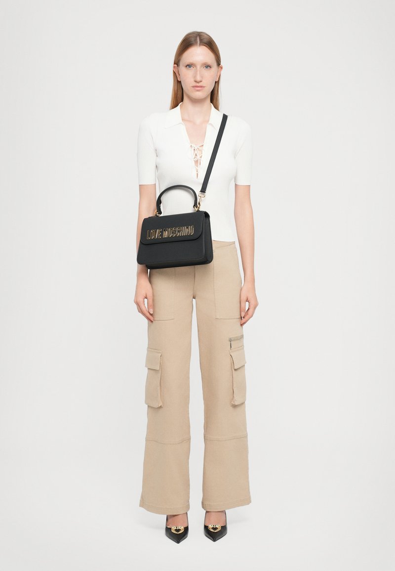 Black handbag with a top handle and shoulder strap, featuring gold "LOVE MOSCHINO" lettering. Model wears a fitted white top and beige cargo pants.