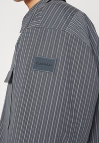 Chemise grise à fines rayures avec une poche poitrine et un patch logo rectangulaire Calvin Klein sur la manche gauche près de l’épaule.