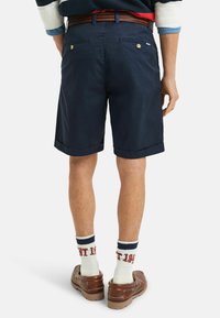 Námornické bavlněné šortky s ohrnutými lemy, dvě zadní kapsy se zapínáním na knoflíky, nošeny s hnědými slip-on botami a bílými ponožkami s červeným textem.