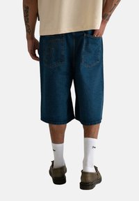 Shorts en denim de couleur bleu foncé, coupe décontractée, dotés de deux poches arrière et d'un logo brodé. Assortis avec des chaussettes blanches et des chaussures brunes.