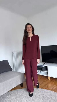 Žena s dlhými tmavými vlasmi, ktorá má na sebe zladený bordový top a nohavice, stojí bosá v modernom obývacom izbe s TV a pohovkou.