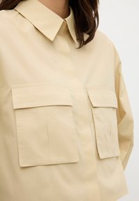 Camicia beige con colletto e bottoni, due tasche anteriori con pattina e vestibilità rilassata. Tessuto morbido e leggero.