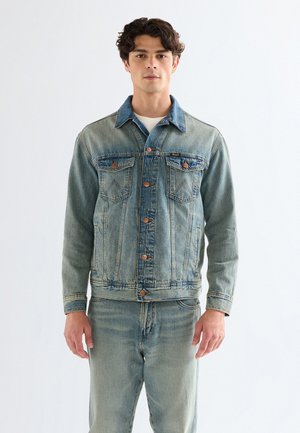 Mladý muž v svetlomodrej rifľovej bunde a džínsoch rovnakej farby, stojaci pred jednofarebným bielym pozadím.