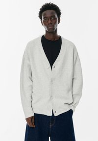 Cardigan grigio chiaro realizzato in morbido tessuto, con scollo a V a costine, bottoni frontali e maniche ampie, abbinato a una maglietta nera e jeans scuri.