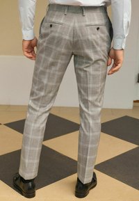 Grau karierte Hose aus einem glatten Stoff. Verfügt über zwei Ges�äßtaschen mit schwarzen Knöpfen und einen taillierten Schnitt. Sichtbare schwarze Schuhe.