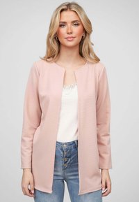 Lichtroze lange mouwen cardigan in een textuurstof met een open voorkant ontwerp. Gedragen over een witte kant tanktop en gecombineerd met blauwe jeans.