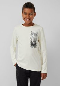 Langärmliges cremefarbenes Baumwoll-T-Shirt mit grafischem Druck in Schwarz und Blau. Rundhalsausschnitt und lockere Passform. Kombiniert mit dunklen Hosen.