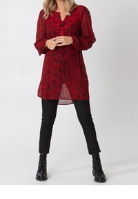Robe tunique à imprimé floral rouge, à manches longues, décolleté en V et texture transparente, associée à un jean skinny noir et des bottes à lacets.
