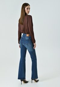 Jeans en denim évasés de couleur bleu moyen, taille haute avec une étiquette en cuir marron, associés à un blouse marron à manches longues et des escarpins noirs.