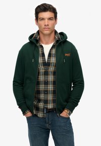 Joven con sudadera con cremallera verde oscuro sobre una camisa de cuadros y jeans, de pie con las manos en los bolsillos frente a un fondo liso.