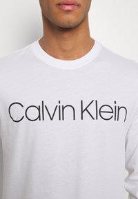 White long-sleeve t-shirt featuring "Calvin Klein" logo in bold black font. Cotton material with a round neckline and smooth texture.