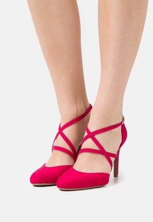 Escarpins en daim rose avec un bout pointu, un talon aiguille fin et des brides croisées à la cheville. Texture lisse avec un matériel minimal.