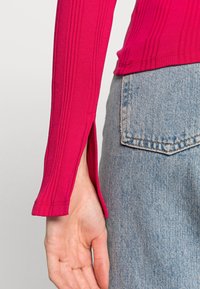 Åtsittande långärmad topp i ljusrosa ribbat tyg, med slitsar på sidorna av fållen, tillsammans med ljusblå utsvängda jeans.