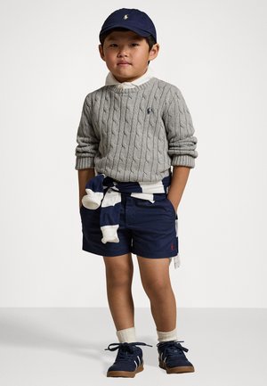 Sweater cinza de ponto trançado, calções azul-marinho com detalhes brancos, meias creme, ténis azul-marinho e boné azul-marinho com logótipo. Suéter padrão amarrado à cintura.