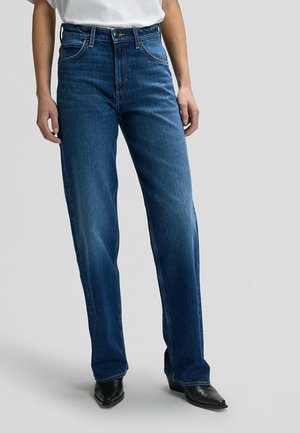 Persoon die donkerblauwe high-waist jeans met rechte pijpen en zwarte laarzen met puntige neus draagt, staand tegen een effen lichtgrijze achtergrond.