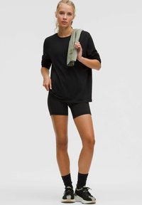 Schwarzes langärmliges Shirt, kombiniert mit schwarzen Radsportshorts. Ein hellgrünes Handtuch über einer Schulter drapiert. Schwarze Sportschuhe mit einer weißen Sohle.