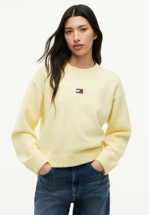 Junge Frau mit dunklen Haaren, die einen hellgelben Pullover und blaue Jeans trägt, steht mit den Händen in den Taschen vor weißem Hintergrund.
