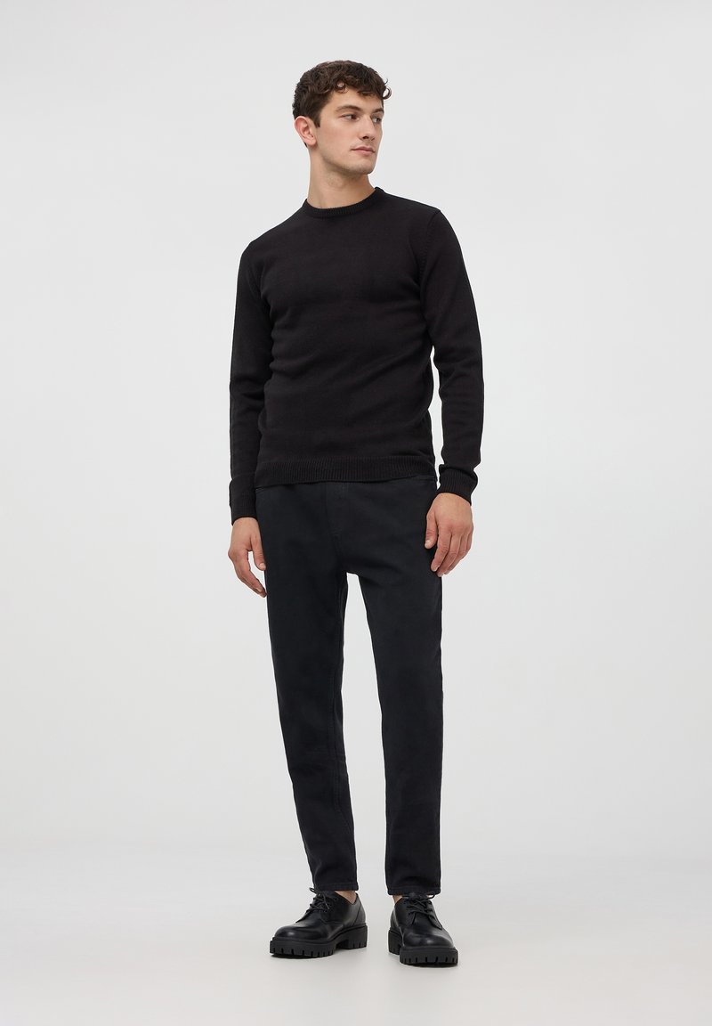 Maglione nero lavorato a maglia con colletto rotondo, abbinato a pantaloni neri slim-fit e scarpe nere con lacci, presentato su uno sfondo neutro.