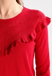 Rode gebreide trui met een gestructureerde kanten inzet met een gerimpelde rand langs de halslijn. Lange mouwen met een nauwsluitende pasvorm.