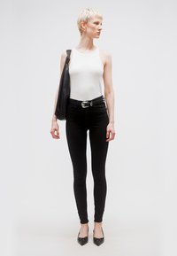 Άτομο με κοντά ξανθά μαλλιά που φορά λευκό αμάνικο τοπ, μαύρα στενά τζιν, μυτερά μαύρα παπούτσια, μαύρη ζώνη και μαύρη τσάντα στον ώμο, κοιτάζοντας πλάγια.