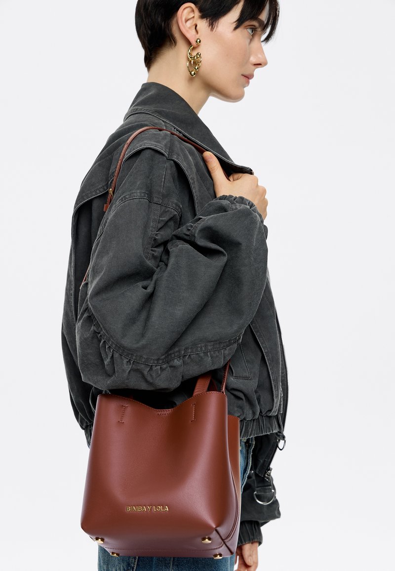 Bruine lederen emmer tas met een gladde textuur, voorzien van gouden hardware en een minimalistischer ontwerp. Draagt aan één schouder met een donkere jas.