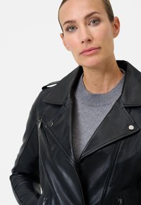 Giubbotto da moto in pelle nera con ampi rever, zip sul petto e tasche, abbinato a un maglione lavorato a maglia grigio sotto.