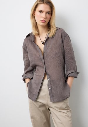 Jeune femme aux cheveux blonds portant une chemise rayée marron, un pantalon beige et un collier pendentif rond noir, posant les mains dans les poches.