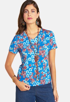Woman wearing a blue floral short-sleeve top, beaded long necklace, large colorful earrings, and blue jeans with hand resting on hip.