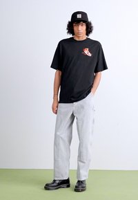 T-shirt noir à manches courtes avec un graphique imprimé rouge et blanc, associé à un pantalon baggy gris clair et des bottes noires.