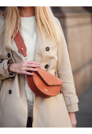 Braune, gebogene Umhängetasche aus glattem Leder mit einem goldenen Logo-Akzent und einem breiten Trageriemen, gehalten von einer Person in einem beigen Trenchcoat.
