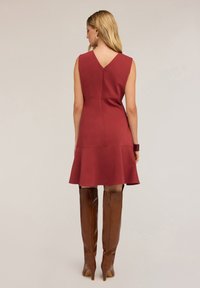 Rotes ärmelloses Kleid mit ausgestellt Saum und V-Ausschnitt auf dem Rücken, gefertigt aus glattem Stoff. Getragen mit braunen kniehohen Stiefeln und einem Armband am Handgelenk.