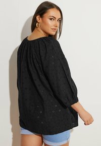 Svart blus med ballongärmar, med ett strukturerat mönster av hexagoner och blommiga detaljer, parat med denimshorts.