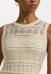 Kvinna som bär en ärmlös krämfärgad stickad topp med spets- och geometriska mönster, samt en guldring i örat.