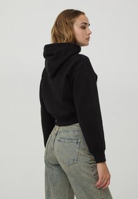Felpa nera corta con cappuccio e cordoncino, realizzata in morbido tessuto di cotone. Abbinata a jeans azzurri a vita alta che mostrano dettagli sulle tasche posteriori.