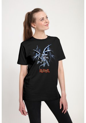 Kvinde med langt hår i en hestehale, iført en sort t-shirt med et blåt drage- og lyn-design, der kigger op og smiler let.
