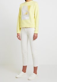 Sudadera amarilla con un estampado gráfico, combinada con jeans blancos recortados y zapatillas blancas, creando un look casual y en capas.