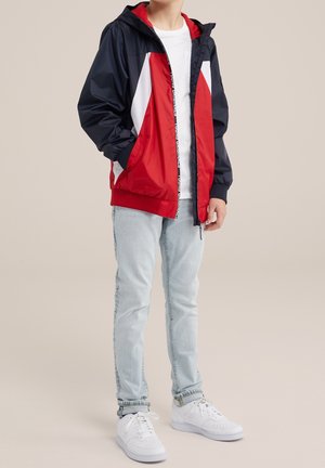 Rød og navy vindjakke med hvide detaljer, lynlåslukning og hætte. Kombineret med lyseblå jeans og hvide sneakers.